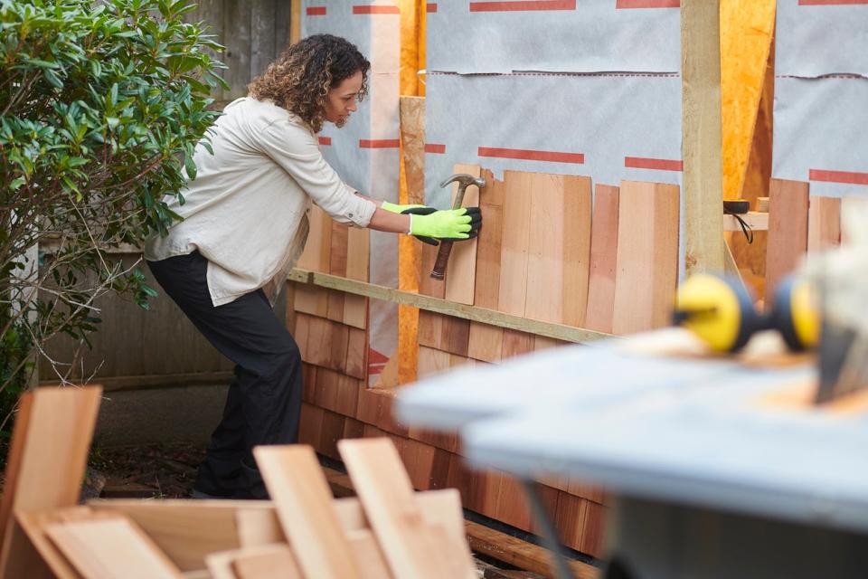 woman-works-on-a-wooden-shed-while-holding-a-hammer-and-wearing-gloves