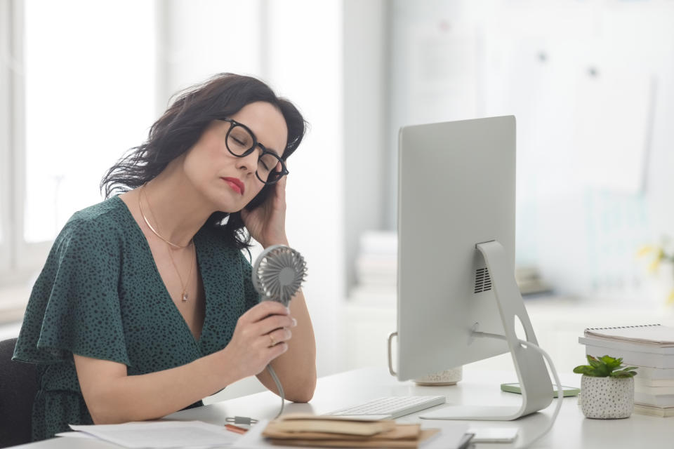 Menopausal mature woman wearing green dress sitting at the desk in the office in front of computer, having hot flashes and using hand fan.