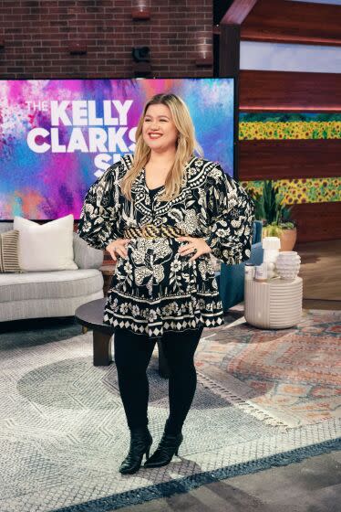 Kelly Clarkson standing with hips on waist, smiling in black and white dress on set of her show