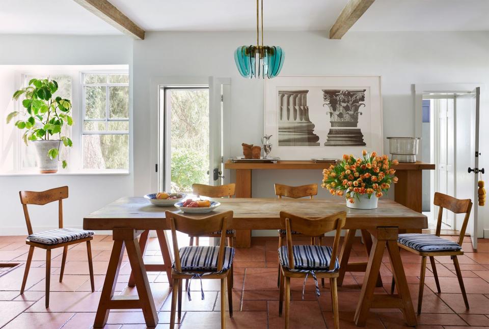 a dining room with a terra cotta tiled floor has a long solid oak table with six wood school chairs with striped cushions, sideboard with architectural prints on the wall, and turquoise glass light pendant