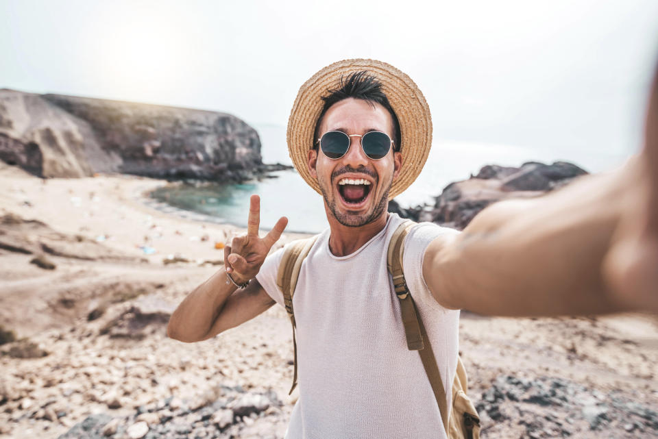 A man in a hat and sunglasses takes a selfie while smiling and making a peace sign on a rocky beach
