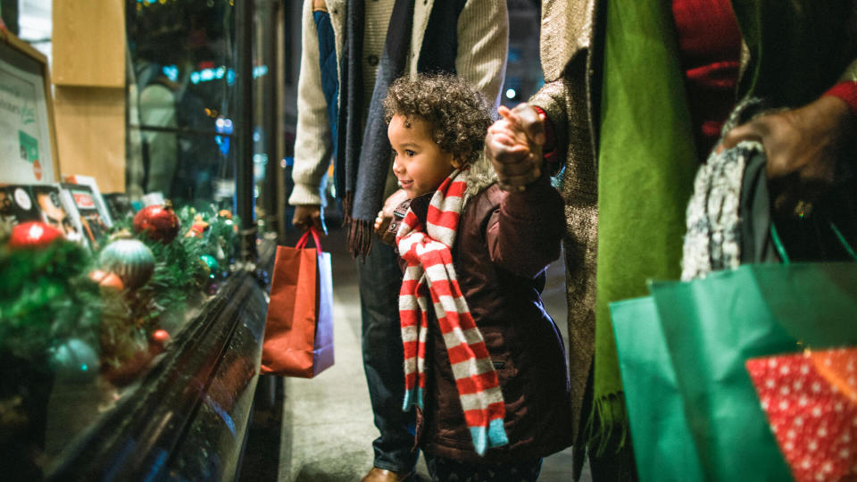 A side-view shot of two grandparents standing outside of a store window with their granddaughter in the city on a cold night, they are wearing warm clothing and holding hands.