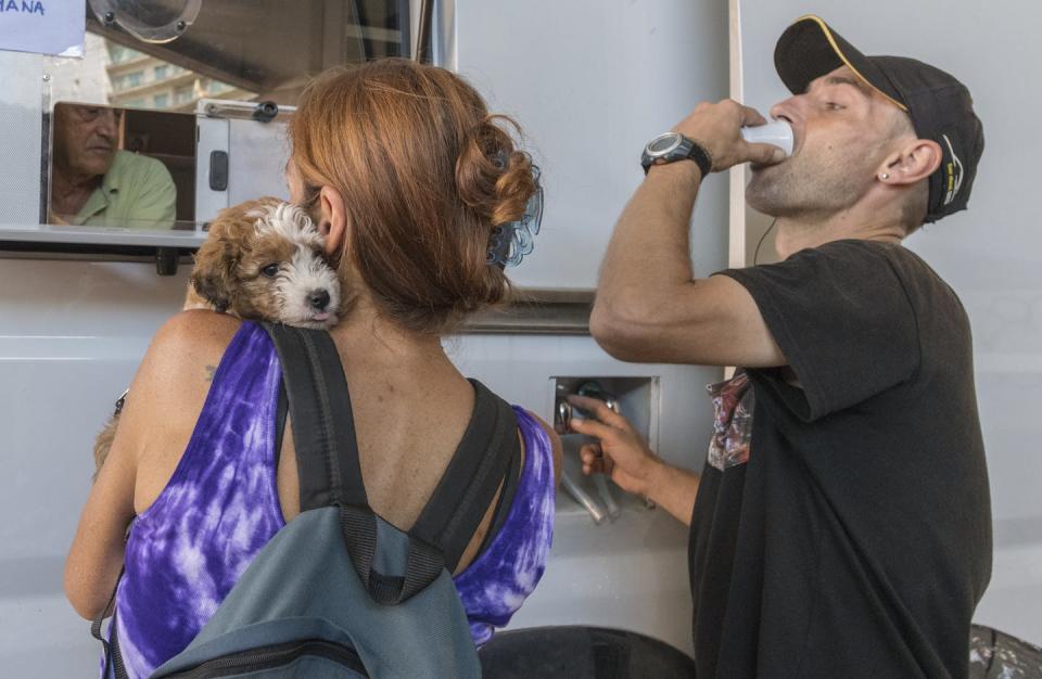 Woman with a dog waits at a white van while a man drinks from a tiny cup