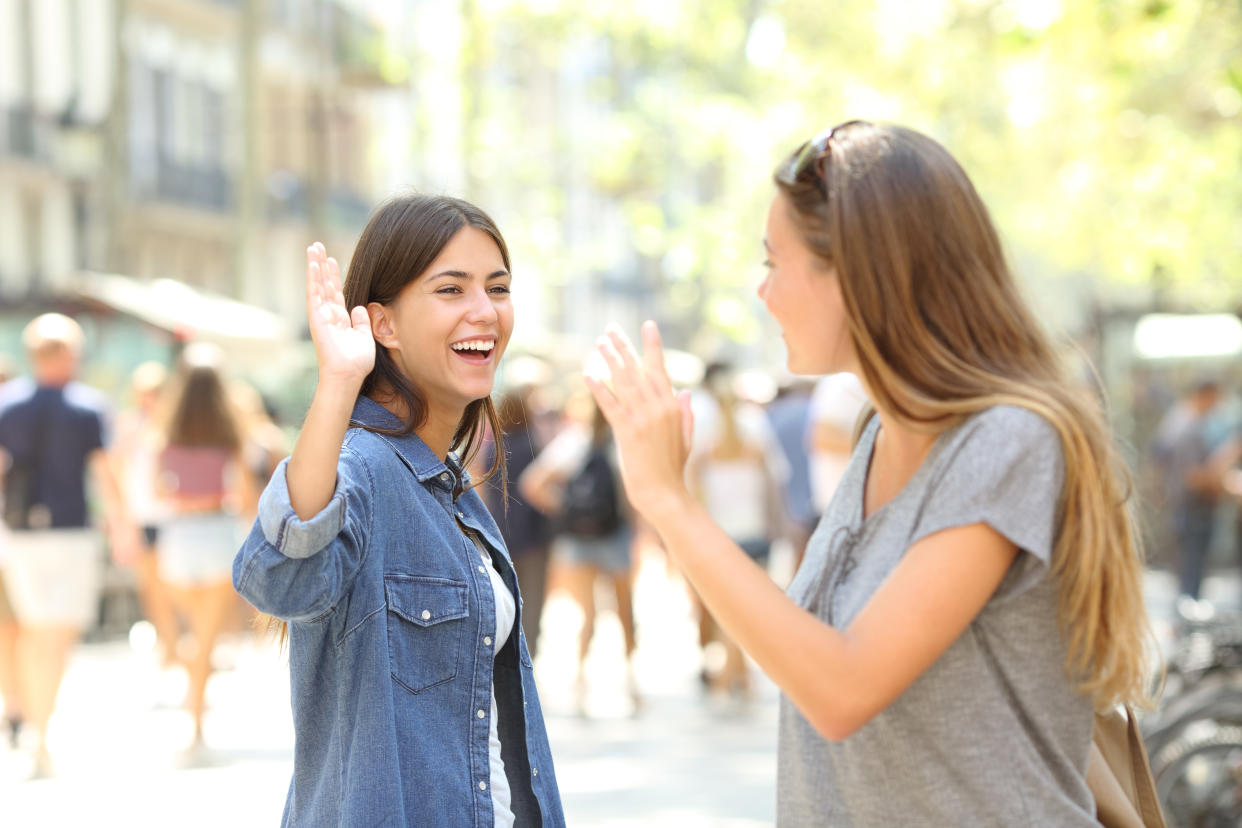 Two young white women passing on a street recognize each other and wave.