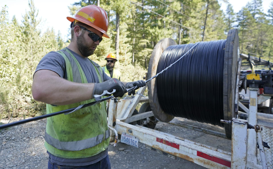 FILE - In this Wednesday, Aug. 4, 2021, file photo, Skylar Core, right, a worker with the Mason County (Wash.) Public Utility District, installs a hanger onto fiber optic cable as it comes off of a spool, while working with a team to install broadband internet service to homes in a rural area surrounding Lake Christine near Belfair, Wash. (AP Photo/Ted S. Warren, File)