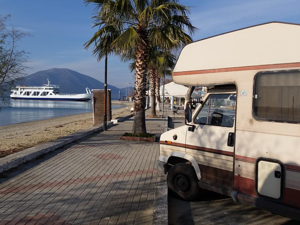 An RV parked next to the water with trees and a boat.