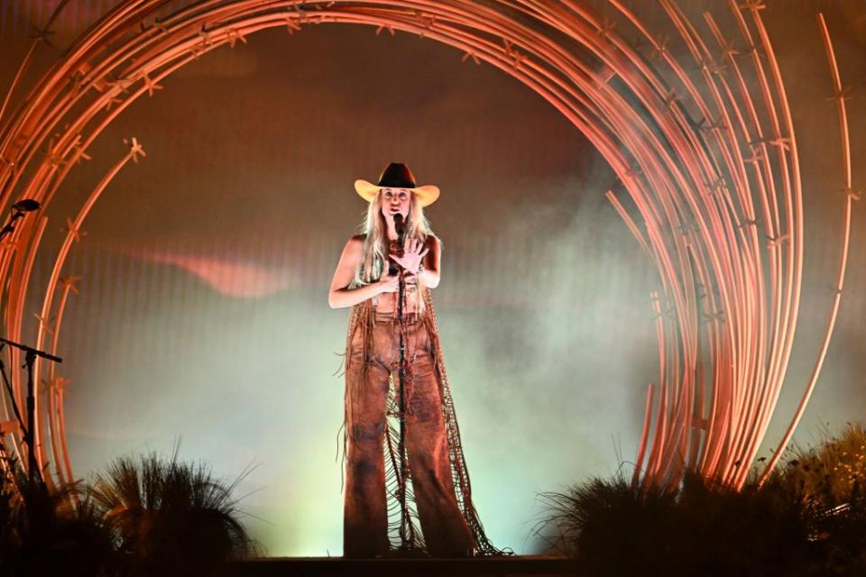 lainey wilson sings into a microphone on a stand and holds a palm out toward the audience with her other hand on her torso, she wears a cowboy hat sleeveless shirt and pants