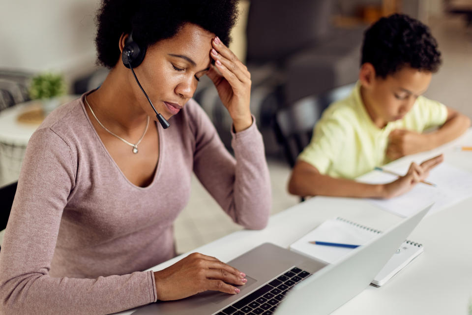 Stay at home mother using laptop and holding her head in pain while her son is doing homework.