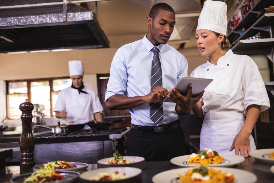 A chef and a waiter in a restaurant kitchen review orders on a tablet computer.