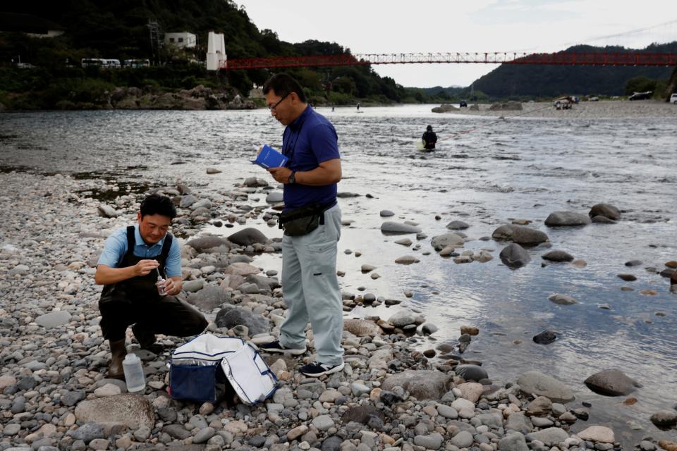 A photo of two men standing on the rocky bank of a river holding a notebook and taking samples of the water.