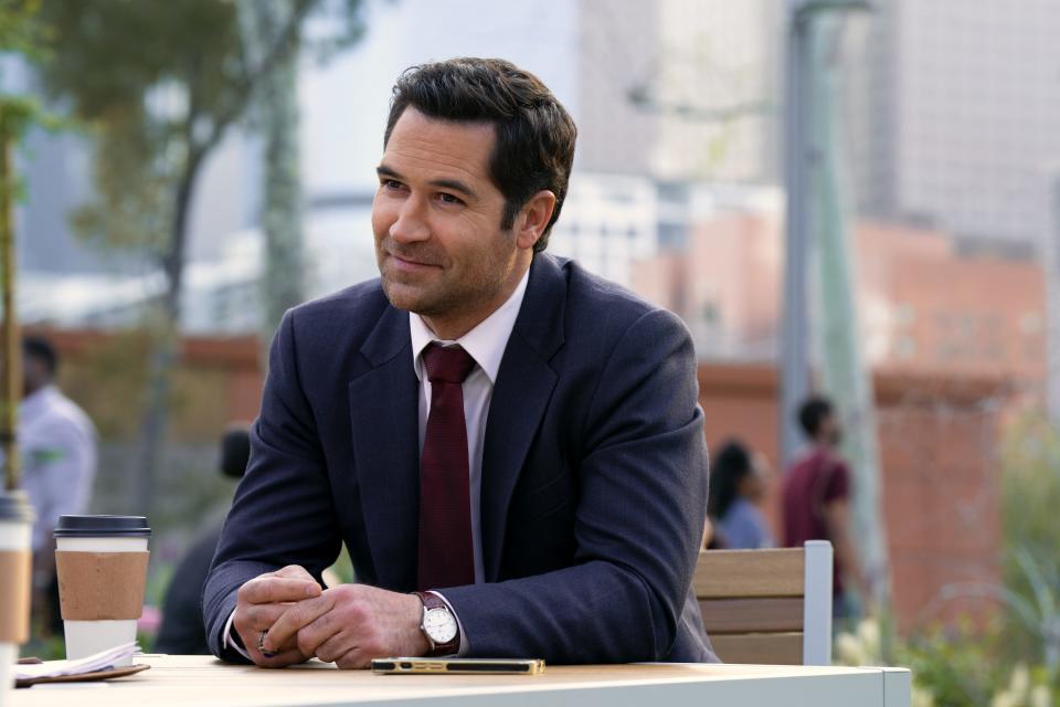 Manuel Garcia-Rulfo in the lincoln lawyer, sitting at a table outside smiling, and wearing a navy suit and red tie