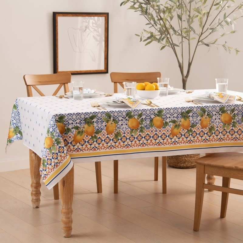 Dining table with a fruit-themed tablecloth and set with dishes and oranges in a bright room