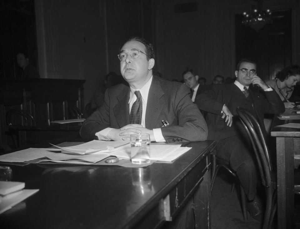 leo szilard wearing a suit and tie, sitting at a table, and speaking to someone off camera