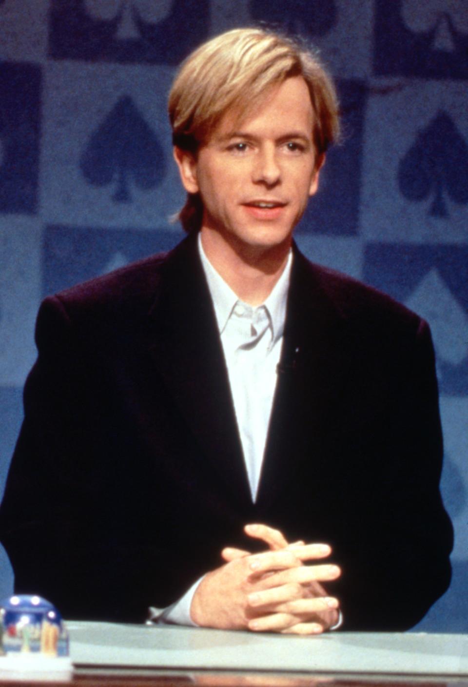 David Spade, seated at a desk with hands folded, wearing a formal suit jacket and shirt, in a setting suggestive of a television show or interview