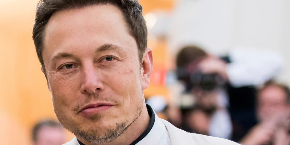 Elon Musk being photographed at a press event while wearing a white suit and black shirt.