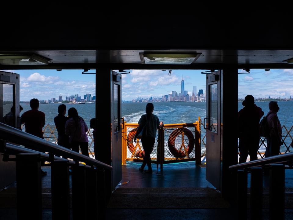 Silhouettes of passengers traveling from New York City aboard the Staten Island Ferry. The Manhattan skyline is seen through the windows in the distance.