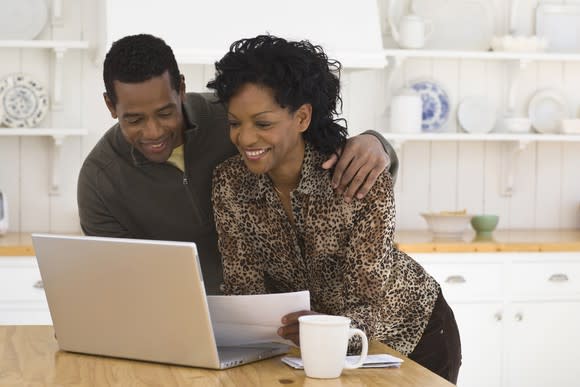 A man and woman smile while looking at a laptop sitting on a table. Also on the table is a coffee cup.