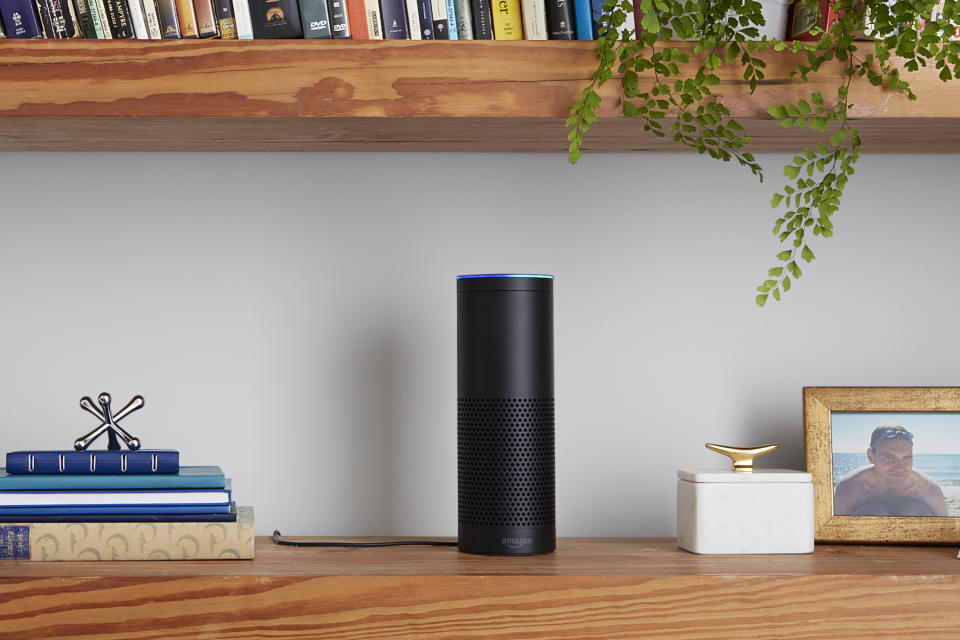 An Amazon Echo speaker on a bookshelf, next to a stack of books and a picture frame