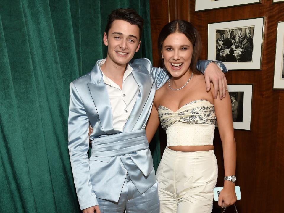 noah schnapp and millie bobby brown at an after party, standing in front of an emerald curtain and brown wall. schnapp is wearing a satin, light blue suit, while brown is in a two piece, strapless white outfit