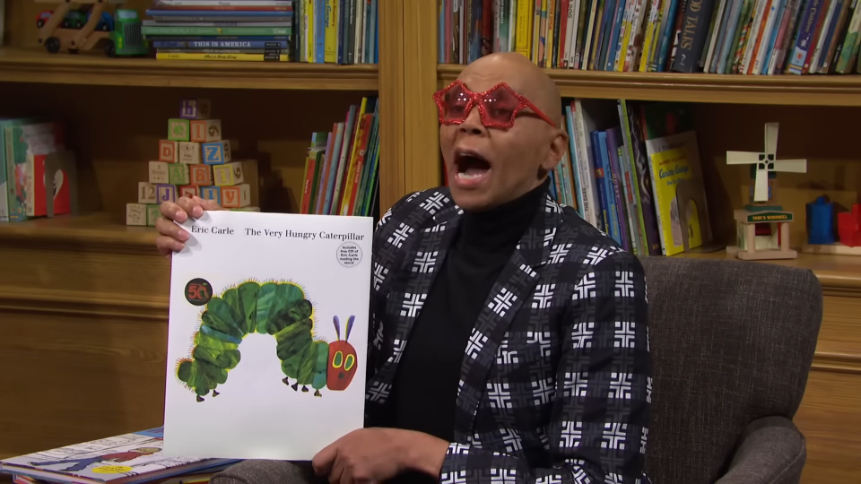 RuPaul, wearing red glasses and a patterned jacket, holds up the book "The Very Hungry Caterpillar" by Eric Carle while enthusiastically speaking