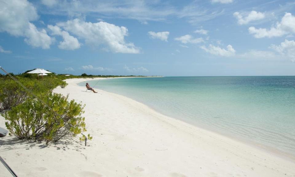 A long empty white sand beach with a man sitting in a deckchair fishing