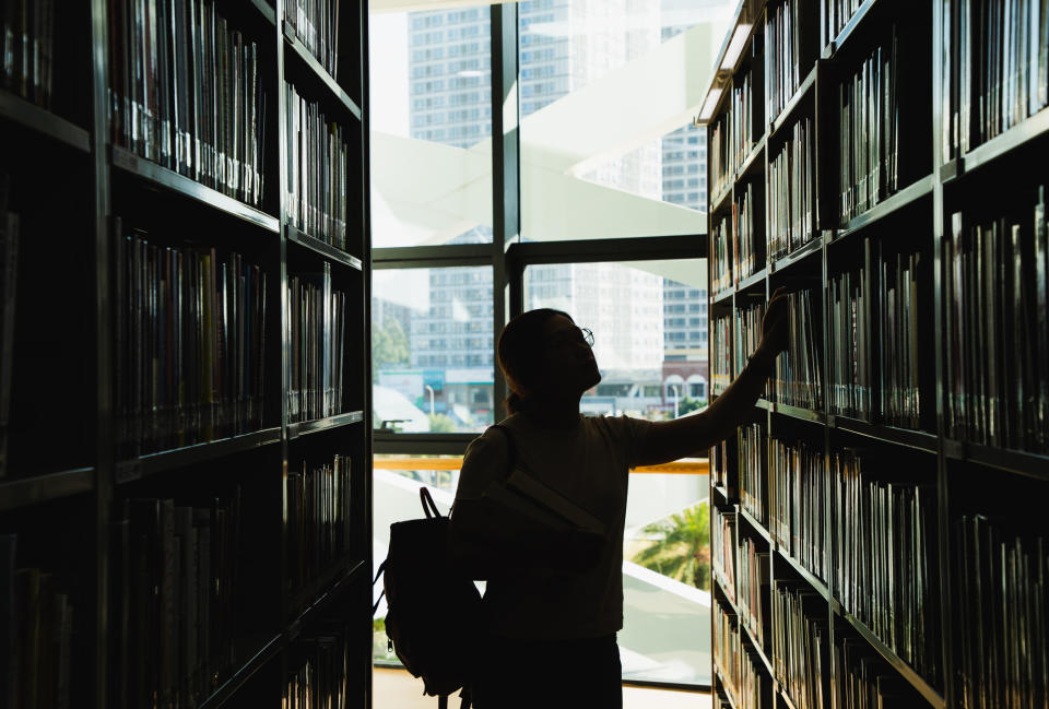 Person in library reaching for a book on a high shelf, cityscape visible through window in background