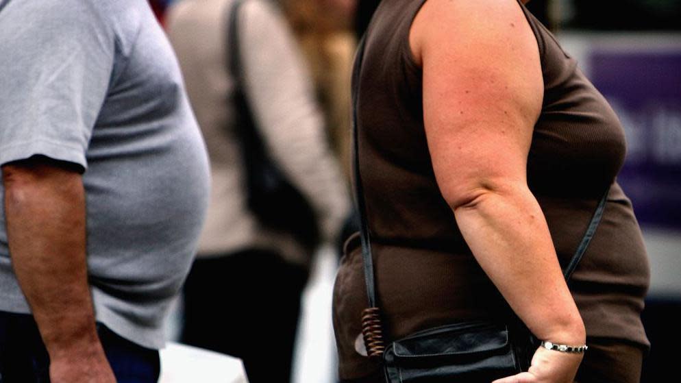 Body shot of a man dressed in a grey t-shirt walking behind a woman dressed in a brown top carrying a handbag