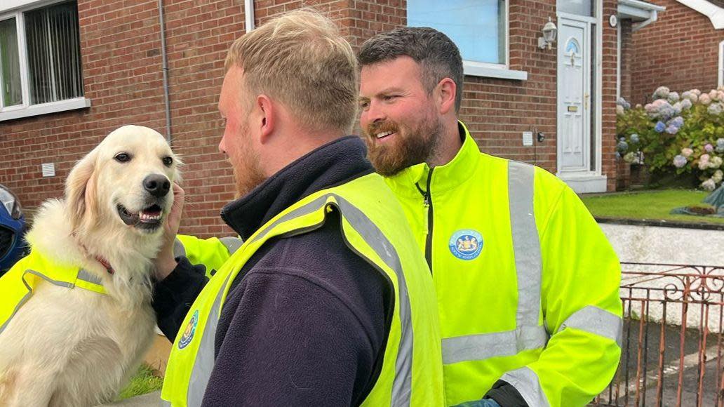 Two men in high vis smile while petting a dog.