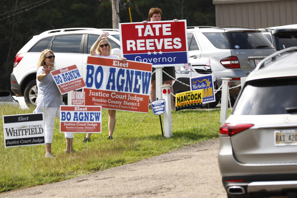 Volunteers from left, Kim Pinnix, left, and her friend Robyn Agnew, center, wife of justice court judge candidate Bo Agnew, wave at incoming voters, while Jake Williams hoists a "Tate Reeves for Governor" sign outside a Flowood, Miss., precinct, Tuesday, Aug. 27, 2019. Runoff candidates of both parties staffed many precincts statewide as they seek runoff wins. (AP Photo/Rogelio V. Solis)