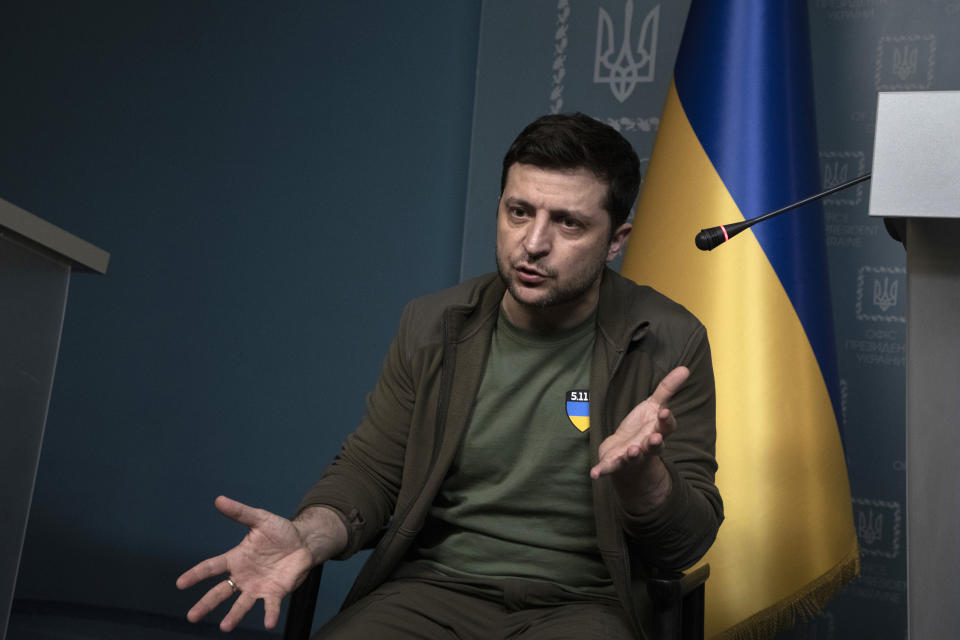 President Volodymyr Zelensky of Ukraine, with a satin Ukrainian flag behind him, and wearing a badge in the same colors, speaks into a microphone.