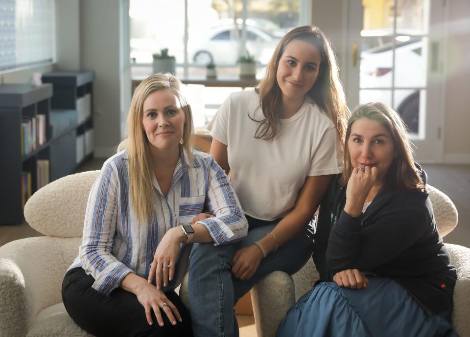 Three women seated on a couch in a store, with a glass window behind them with a view to the street.