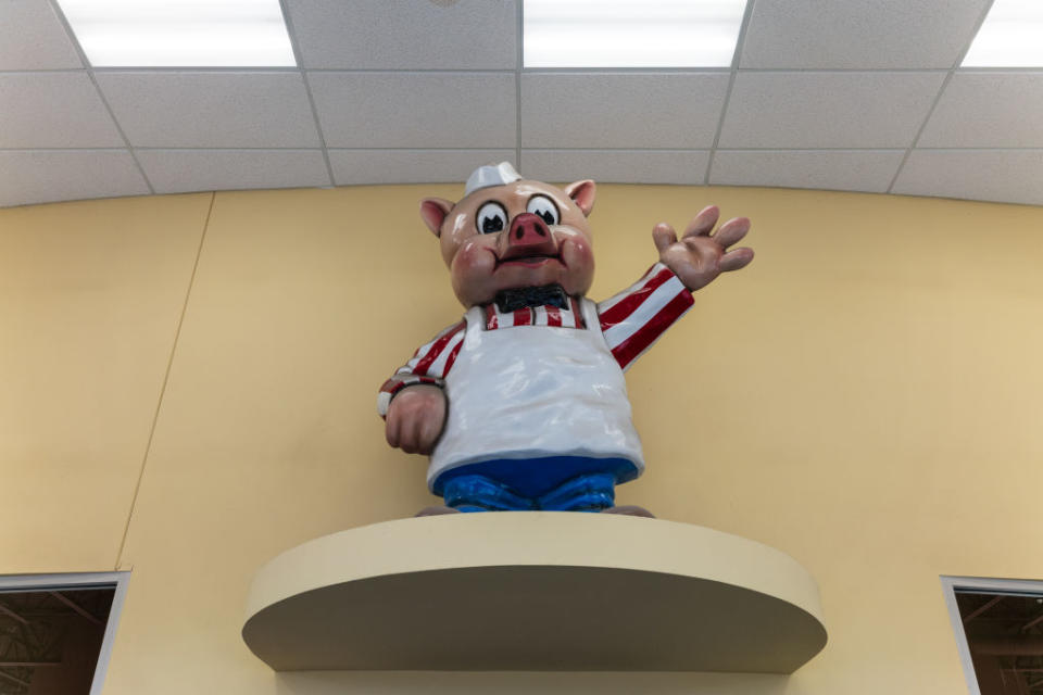 A statue of a pig wearing a chef's hat, a striped shirt, an apron, and blue pants, standing on a platform and waving