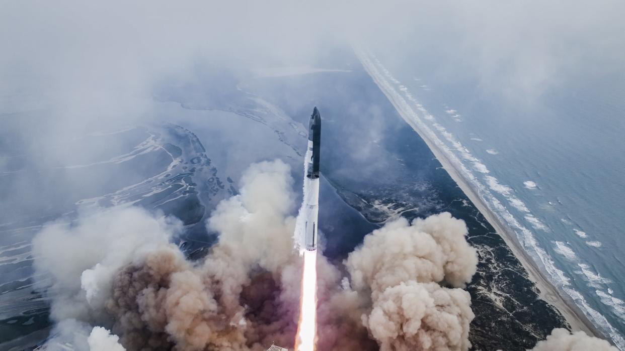  A large silver rocket flies through a blue sky above a column of flame. 