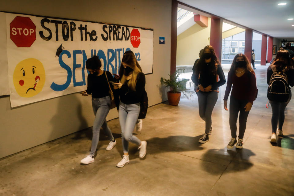 Students wearing face masks walk along a school hallway beneath a sign that reads: Stop the spread. Senioritis 2022.