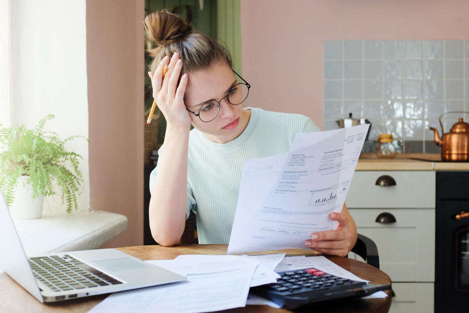 Indoor shot of young European Caucasian girl looking at financial documents at home with deeply bored face looking sick and tired of her economic problems, trying to check counts and all details