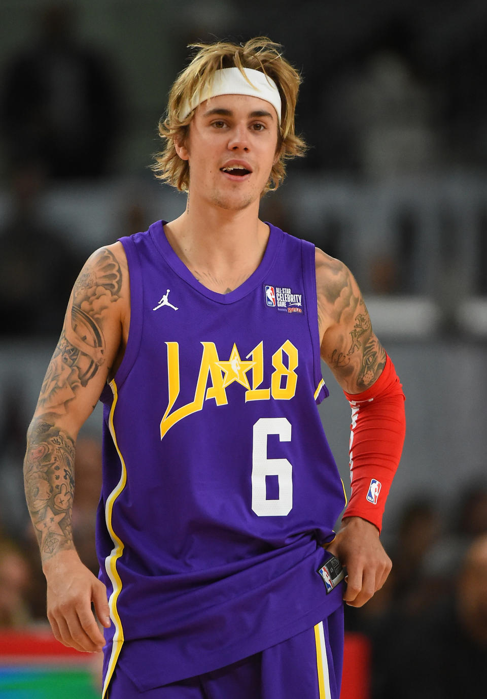 Justin Bieber wearing a purple basketball jersey with "LA 18" and the number 6, a white headband, and a red arm sleeve, standing on a basketball court