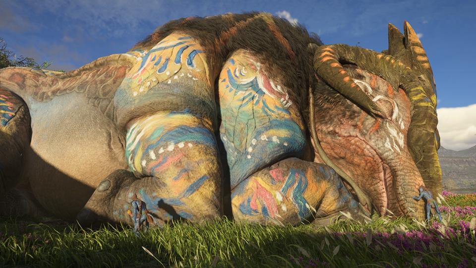 Still from the video game Avatar: Frontiers of Pandora. An enormous land beast (around the size of blue whale) with six limbs, ram-like curly horns, brown fur around it's neck and intricate blue and white markings drawn on it is lying on its side in a deep sleep. Two humans (very tiny in comparison) are inspecting the large beast.