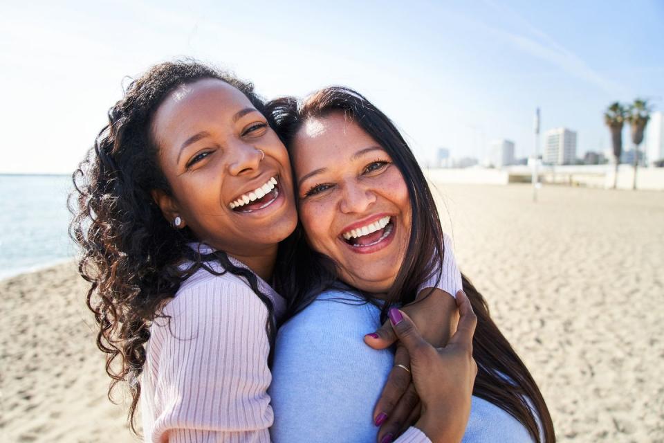 close up photography of multiracial family, looking and smiling at camera, daughter hugging her mother, standing on the beach, woth big smile