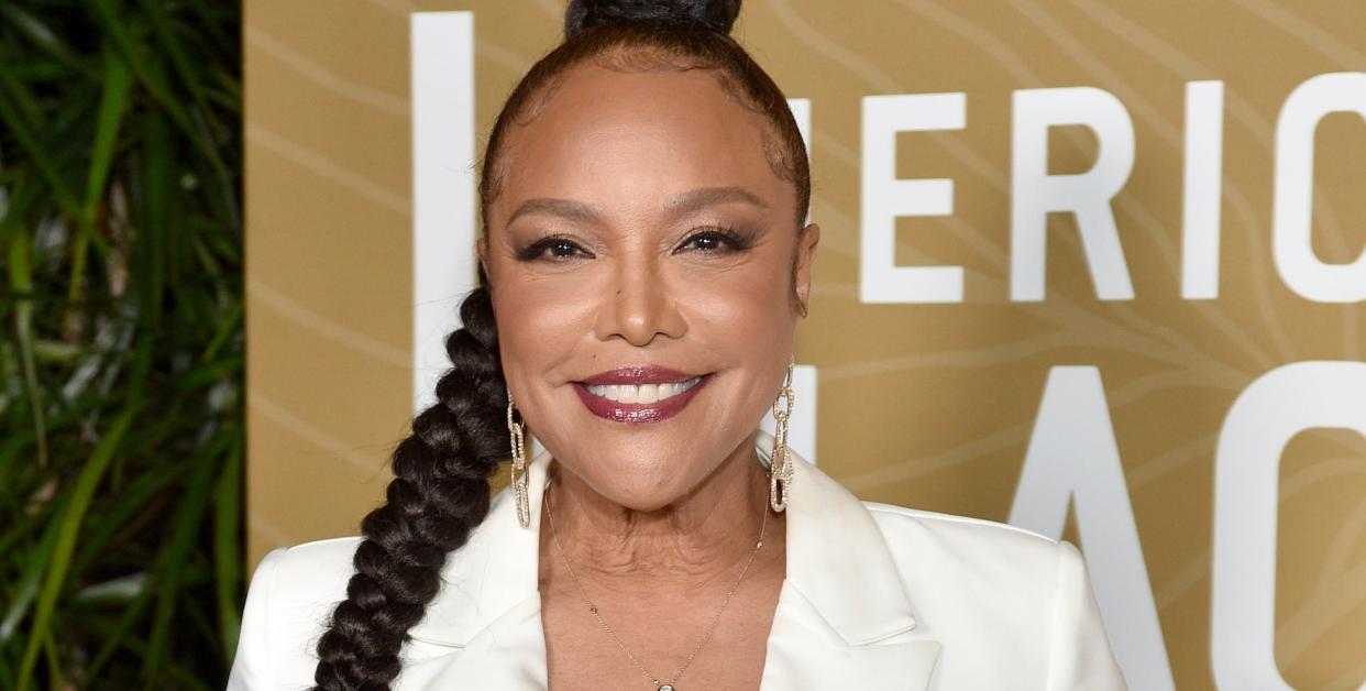 lynn whitfield, a woman stands looking at the camera and smiling, she wears her long black hair in a braid and a white suit top and trousers