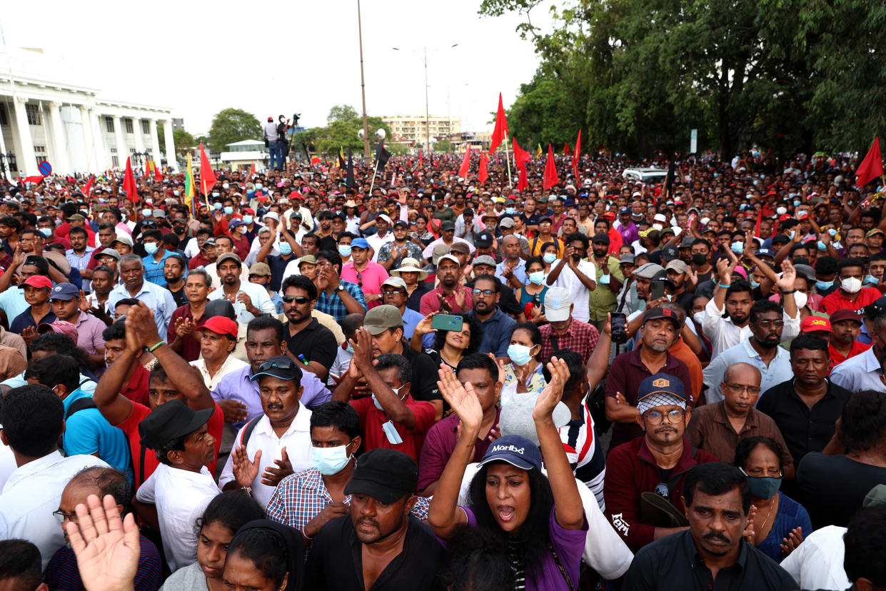 Demonstrators of People's Liberation Front, many holding red flags, cheer at a packed protest of hundreds of people, with a white building with columns in the background..