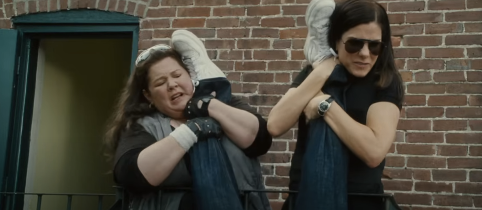 Melissa McCarthy and Sandra Bullock balance on a railing, each holding one leg up near their heads, appearing to be in a challenging or comedic situation