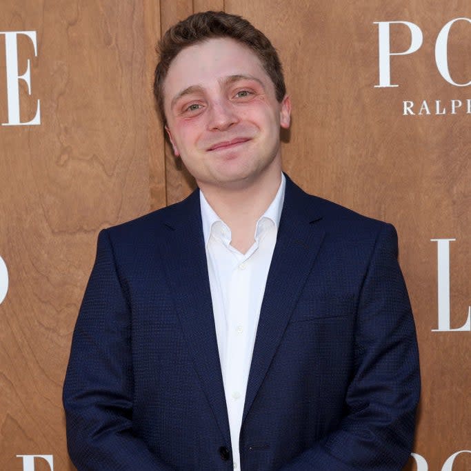 A man in a suit stands in front of a wooden backdrop featuring the text 