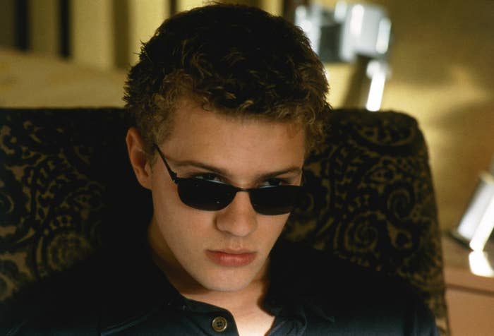 A young man with short curly hair is wearing dark sunglasses and a serious expression. He is sitting on a patterned chair