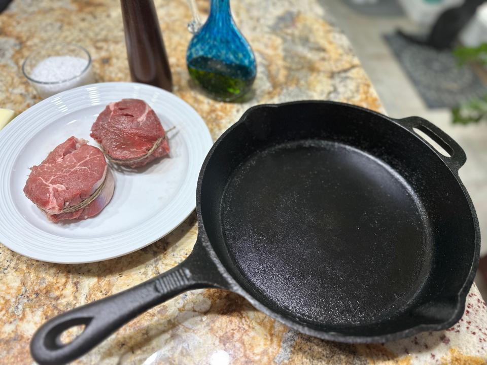 Two raw filet mignon steaks with brown kitchen twine tied around the sides next to a cast iron pan on a brown stone countertop