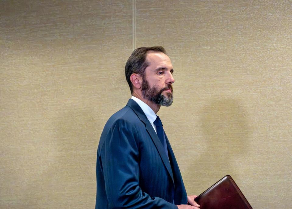 Jack Smith, framed waist and up, walks in front of a sand-color wall while holding a leather documents holder