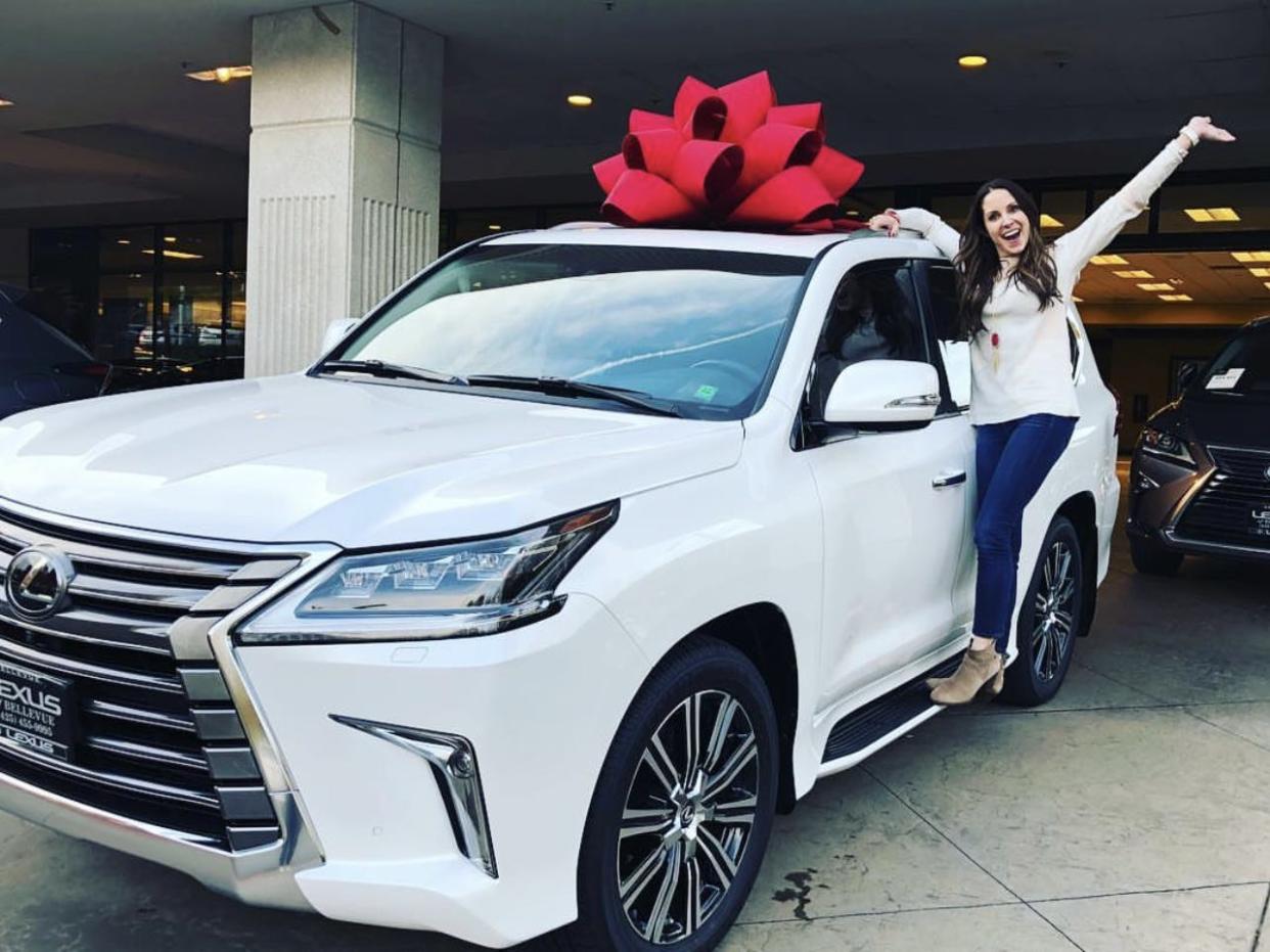 A joyful woman stands on the side of a Lexus with a red bow on its roof