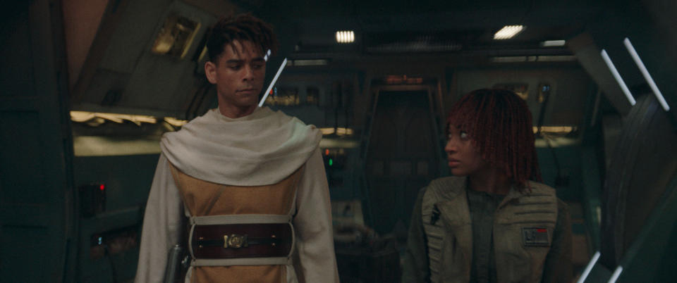 Jay Lycurgo and Ellora Torchia in a futuristic spaceship set, Jay in a robe with a belt, and Ellora in a tactical outfit looking at each other