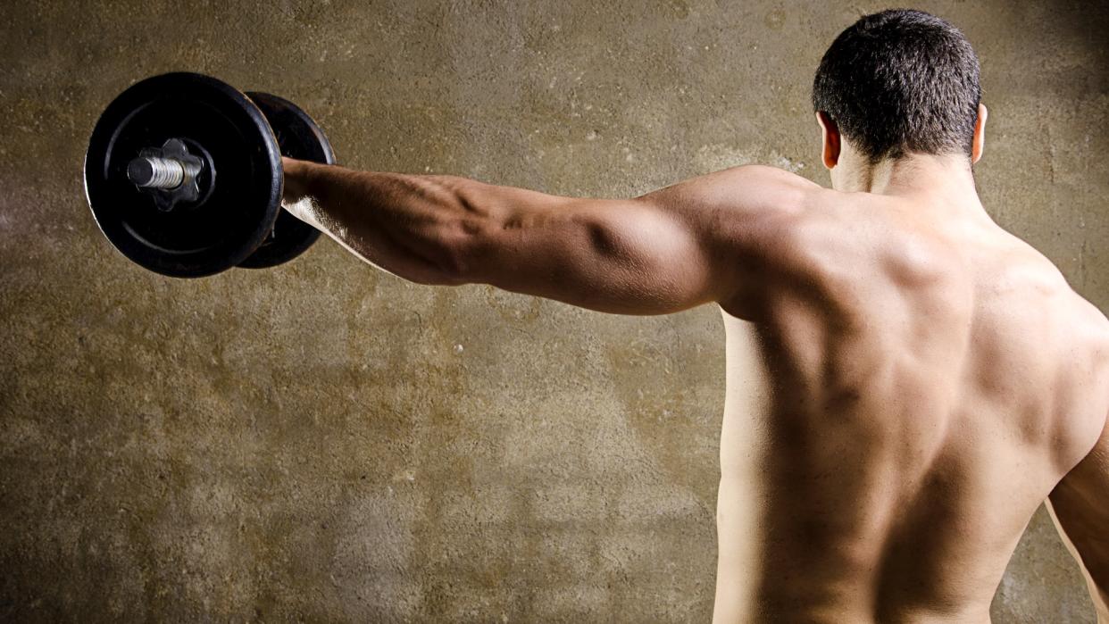  Man performing a lateral raise exercise using a dumbbell in his left hand with back to camera 