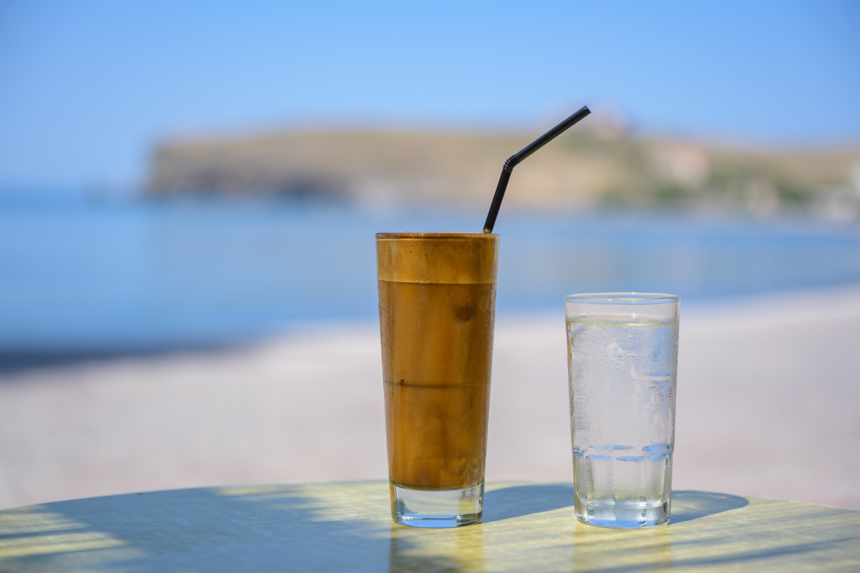 A glass of Greek Frappe Coffee and a glass of water, on a table by the sea, Lesvos, Greece, beach, sea, and sky blurred in the background