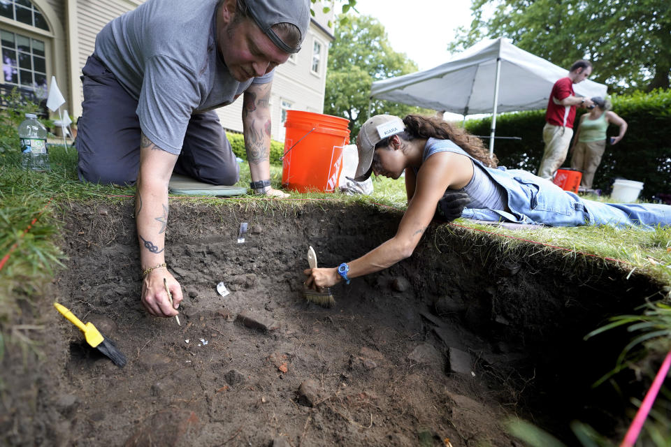 University of Massachusetts Boston graduate students Nicholas Densley, of Missoula, Mont., left, and Kiara Montes, of Boston, right, use brushes while searching for artifacts at an excavation site, Wednesday, June 9, 2021, on Cole's Hill, in Plymouth, Mass. The archaeologists are part of a team excavating the grassy hilltop that overlooks iconic Plymouth Rock one last time before a historical park is built on the site. (AP Photo/Steven Senne)